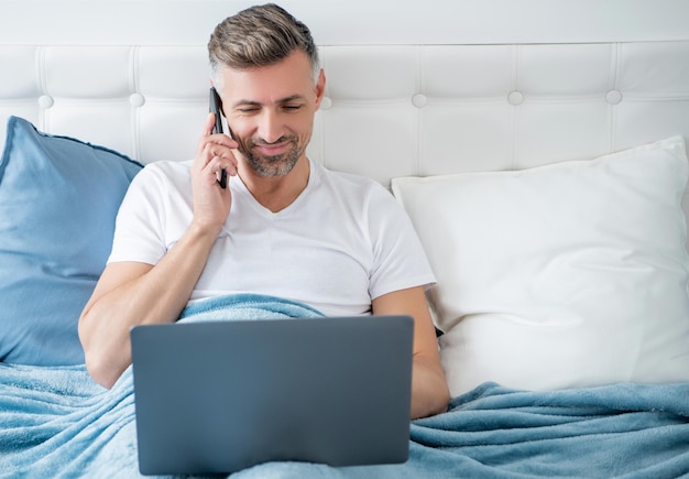 Mature man speaking on phone and working on laptop in bed