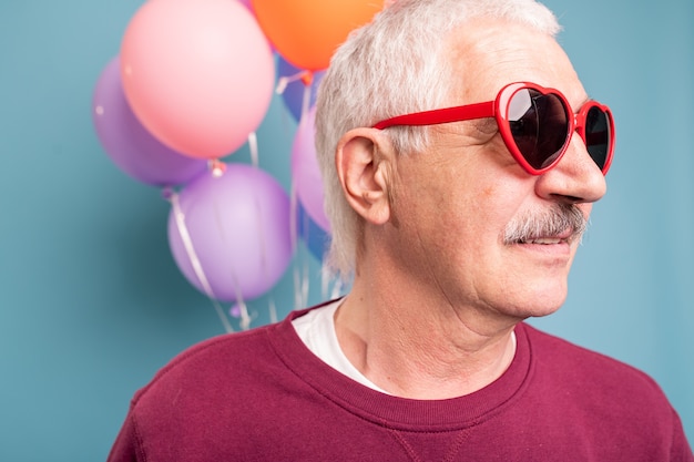 Mature man in stylish sunglasses posing on blue wall with colorful balloons
