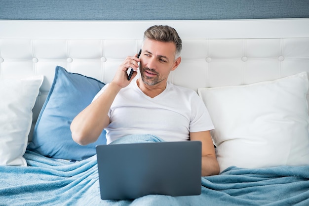 Mature man talk on phone and working on laptop in bed