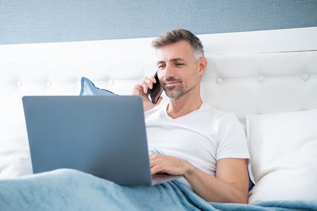 Mature man talking on phone and working on laptop in bed