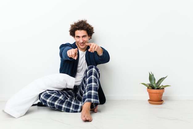 Mature man wearing pajama sitting on house floor cheerful smiles pointing to front.