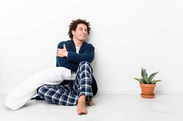 Photo mature man wearing pajama sitting on house floor hugs, smiling carefree and happy.