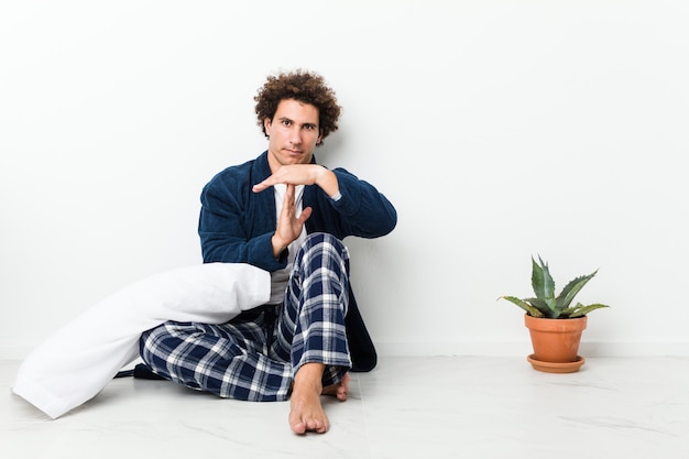 Mature man wearing pajama sitting on house floor showing a timeout gesture.