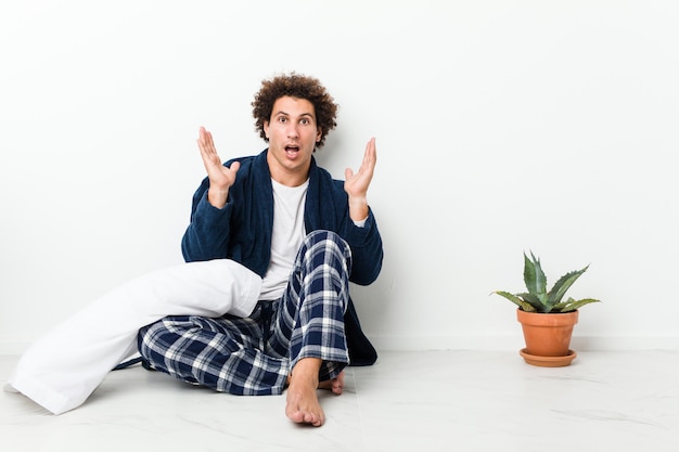 Mature man wearing pajama sitting on house floor surprised and shocked.