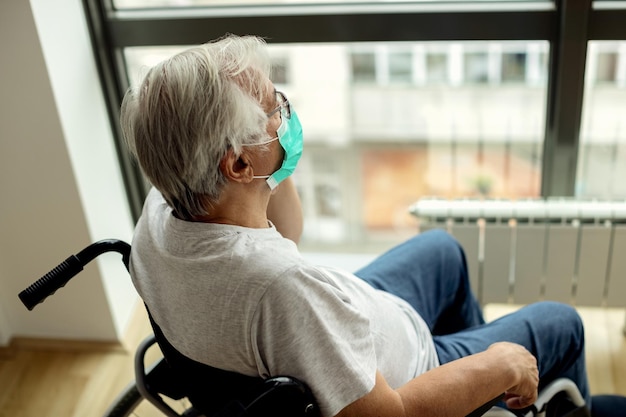 Mature man in wheelchair looking through the window during coronavirus lockdown