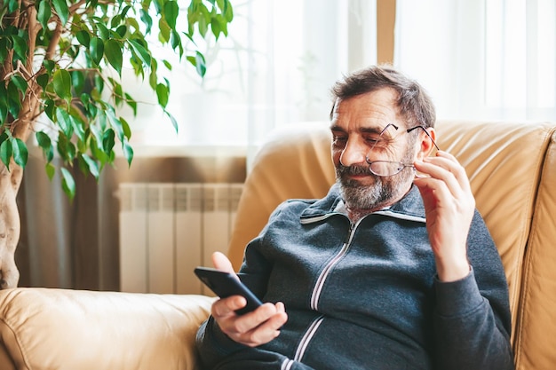 ソファに座ってスマートフォンで何かを検索または読んでいる眼鏡をかけた成熟した男性テクノロジーコンセプトを使用して高齢者