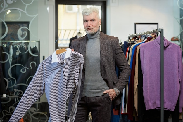 A mature man with gray hair is posing and showing a shirt in a clothing store. A male customer with a beard wears a wool suit in a boutique.