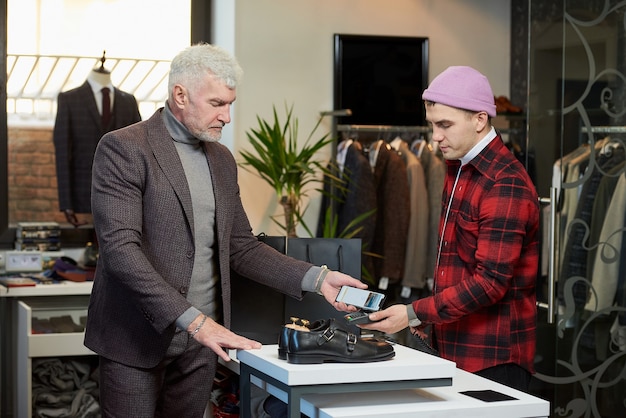 Un uomo maturo con i capelli grigi e un fisico sportivo sta applicando un cellulare a un terminale di un punto vendita in un negozio di abbigliamento. un cliente maschio con la barba paga un commesso in una boutique.