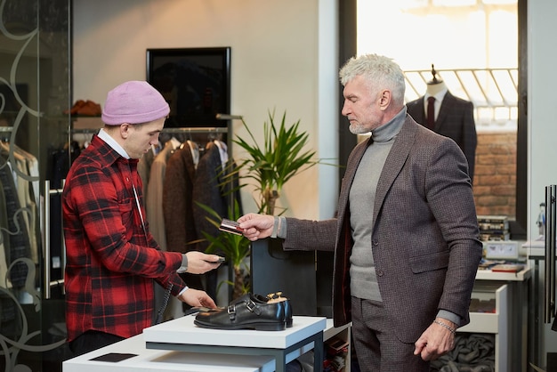 A mature man with gray hair and a sporty physique is handing a credit card to a seller to pay for purchases in a clothing store A male customer with a beard and a shop assistant in a boutique