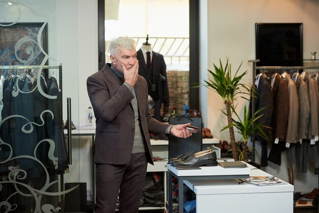 Foto un uomo maturo con i capelli grigi e un fisico sportivo ha in mano una carta di credito per pagare gli acquisti in un negozio di abbigliamento. un cliente con la barba si sta massaggiando il viso in una boutique