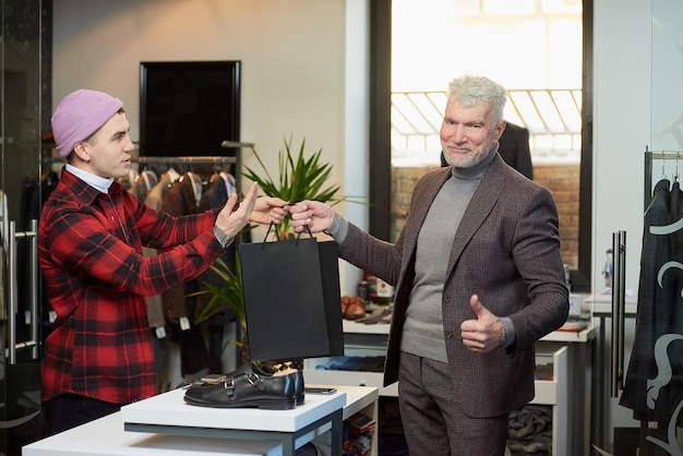 A mature man with gray hair and a sporty physique is taking black paper bags from a seller and showing thumbs up in a clothing store A shop assistant is giving paper bags to a male customer