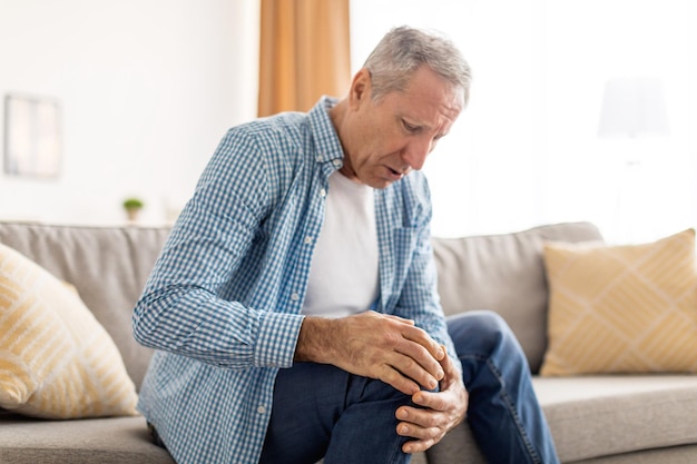 Mature man with knee pain sitting on couch at home