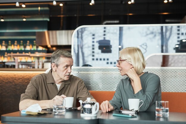 Mature man with mustache sitting at table and telling story to curious girlfriend while they drinking tea in cafe