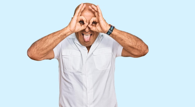 Mature middle east man with mustache wearing casual white shirt doing ok gesture like binoculars sticking tongue out eyes looking through fingers crazy expression