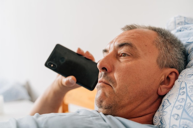 Uomo maturo di mezza età che ascolta un messaggio audio e sdraiato su un letto anni '50 per il tempo libero con la tecnologia