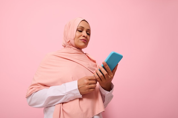 Photo mature muslim woman of middle eastern ethnicity in pink hijab with smartphone in blue cover on her hands, isolated on pink pastel background with copy space