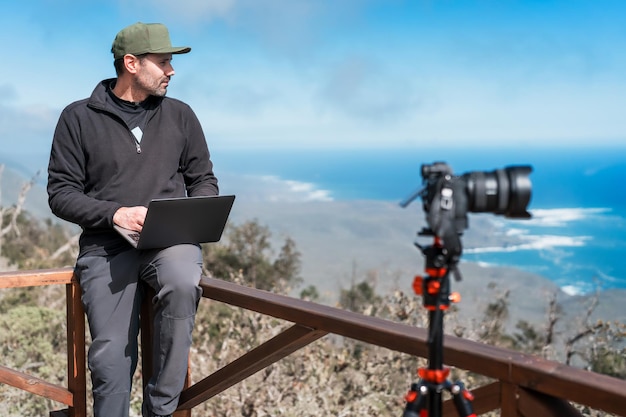 Зрелый человек с ноутбуком и фотоаппаратом, сидящий на смотровой площадке и работающий над портретом Тихого океана