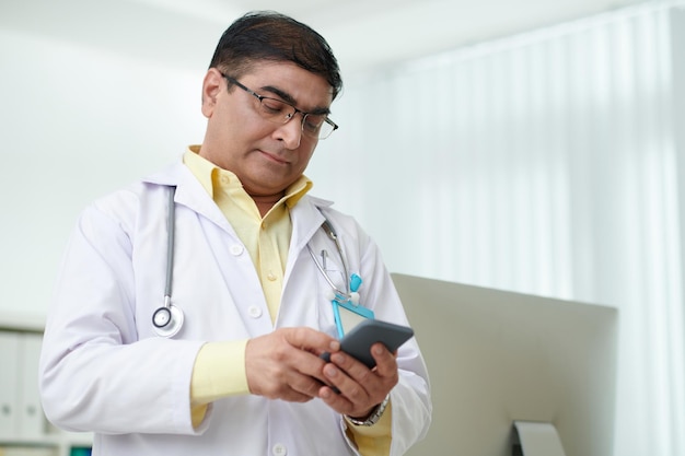 Medico maturo in piedi in studio medico e che controlla messaggi di testo o notifiche sullo smartphone