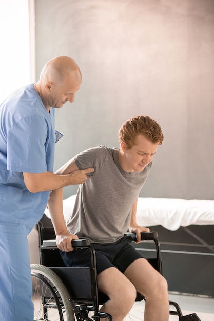 Mature physiotherapist helping sick young man to sit in wheelchair after rehabilitation training while supporting his arm and elbow