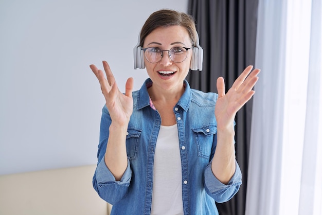 Mature positive talking middle aged woman in headphones looking at camera. Female working remotely, communicating with business colleagues and partners. Woman talking family friends while traveling