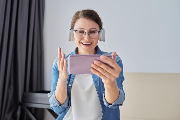 Mature positive talking woman in headphones looking at smartphone screen, video call. Working remotely, communicating with business colleagues. Woman talking to family and friends on vacation travel