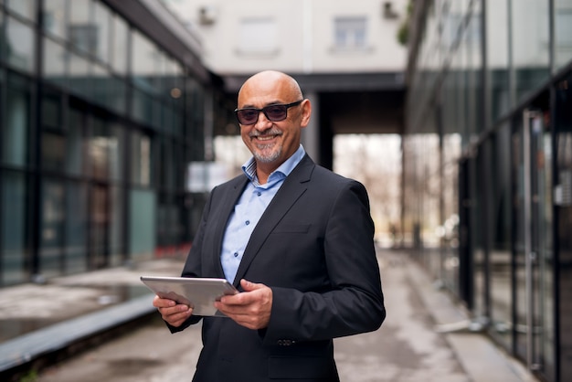 Mature professional elegant businessman is holding tablet while standing in the passage of narrow street.