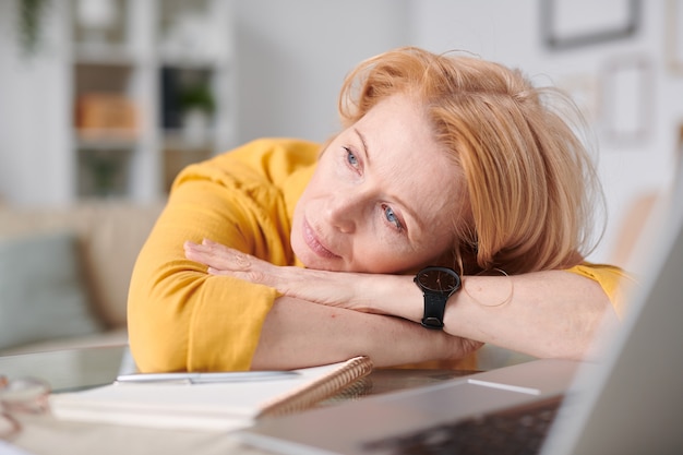 Matura imprenditrice rilassata con i capelli biondi mantenendo la testa sulla scrivania mentre pensa a qualcosa di piacevole in pausa durante il lavoro a distanza