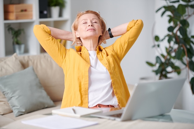 Photo mature restful blond woman in casualwear keeping her hands by backhead while sitting by workplace at home during self isolation period