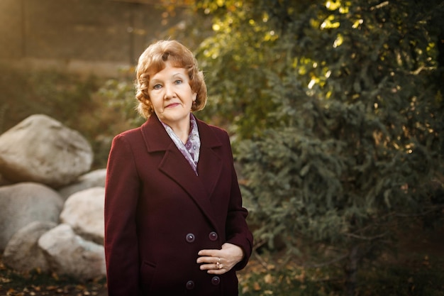 A mature russian woman with short hair stands in the forest in a red buttoned coat