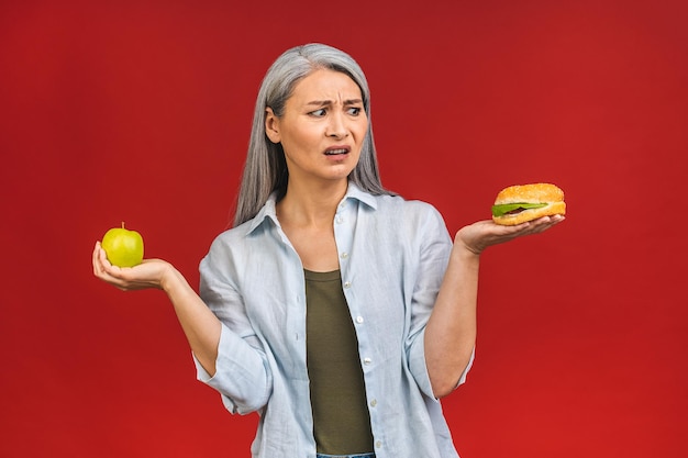 Photo mature senior asian woman holding burger and green apple in hands stop sign to unhealthy food isolated over red background