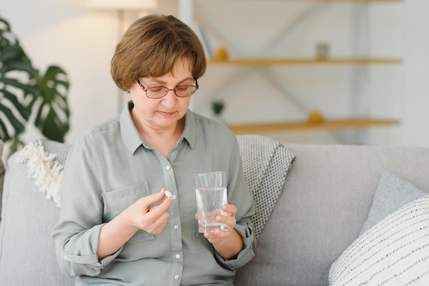 Зрелая пожилая женщина средних лет держит таблетку и стакан воды, принимая обезболивающее, чтобы облегчить боль, лекарства, витамины, антибиотики, лекарства для пожилых людей, концепция, вид крупным планом