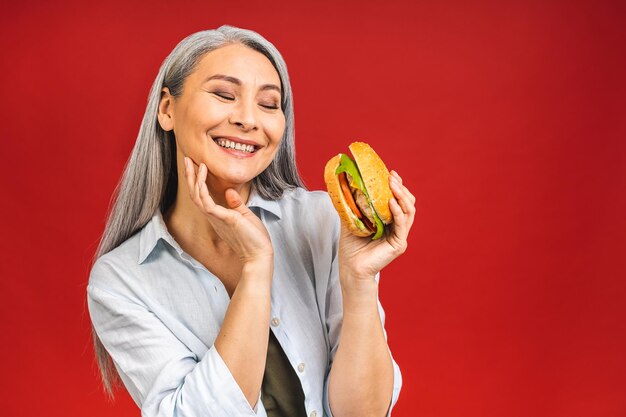 満足のいくハンバーガーを食べる成熟した年配の女性おばあちゃんは、赤い背景の上に孤立して空腹の立っている間、おいしいハンバーガーテイクアウトハンバーガー注文ファーストフード配達のおいしい一口を楽しんでいます