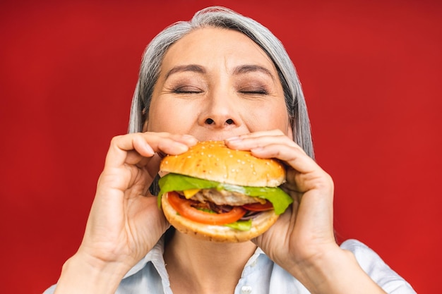 만족스럽게 햄버거를 먹는 성숙한 할머니 할머니는 빨간 배경 위에 고립된 배고픈 동안 맛있는 햄버거 테이크아웃 햄버거 주문 패스트푸드 배달을 즐깁니다.