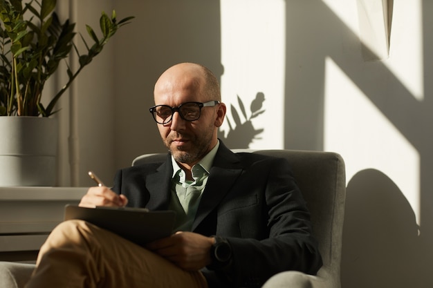 Mature serious businessman in eyeglasses sitting on armchair and making notes in document he working at office