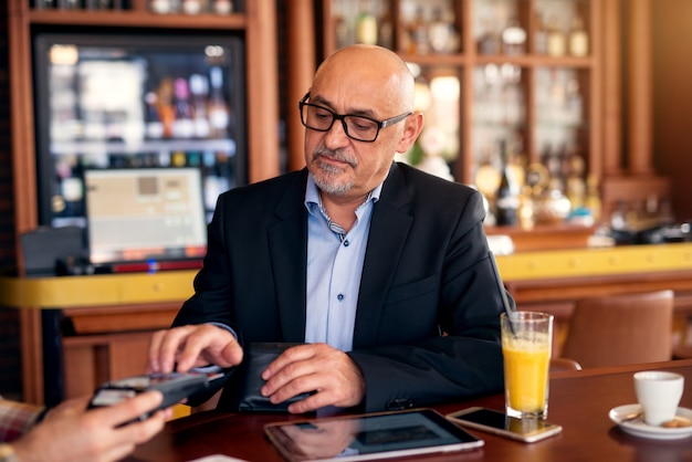 Mature serious professional businessman is using a tablet and paying check with the credit card while sitting in a coffee shop.