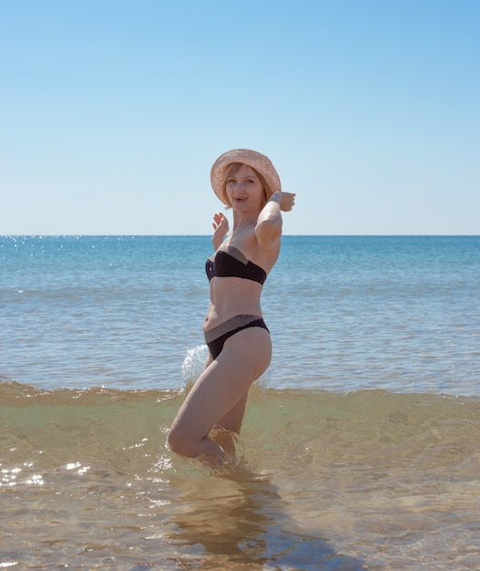 A mature slender woman in a swimsuit and a hat laughs in the sea waves. Sea vacation and travel.