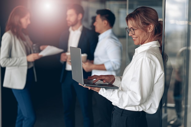 Photo mature smiling businesswoman at office with group of colleagues on background working on laptop