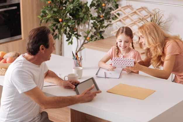 Mature smiling handsome father enjoying family holiday at home and using tablet while wife giving present to the daughter