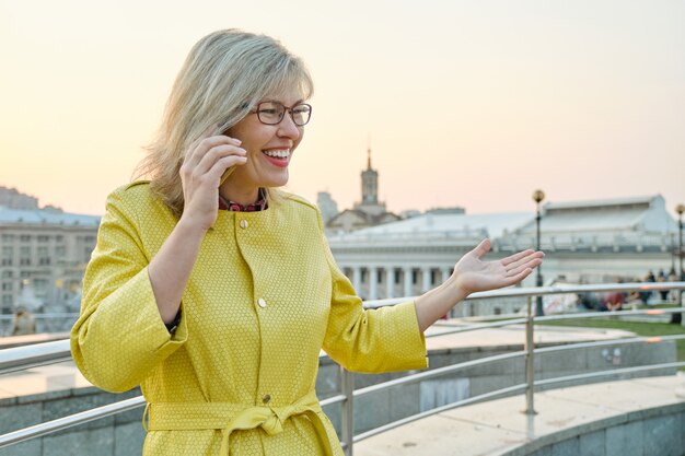 Mature smiling woman in glasses, yellow coat talking on mobile phone