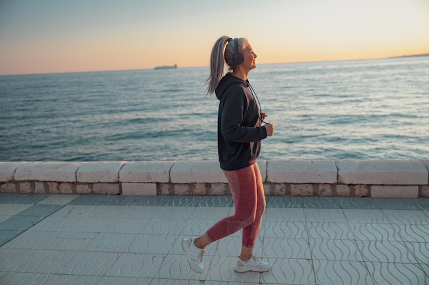 Mature sportwoman wearing headphones and sports clothing jogging on the embankment