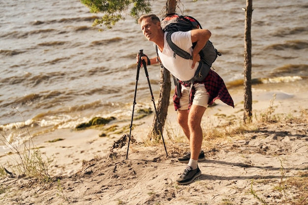 Зрелый спортивный мужчина занимается скандинавской ходьбой у берега моря и по дороге страдает от боли в пояснице