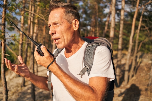 Mature sporty male is walking in forest with tourist rucksack and communicating via radio connection