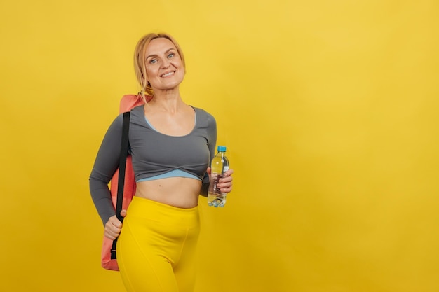 Mature sporty woman holding yoga mat and bottle of water on yellow background