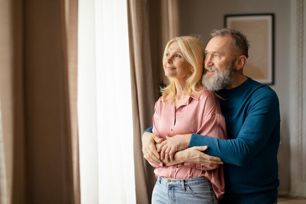 Mature spouses embracing looking aside standing near window at home