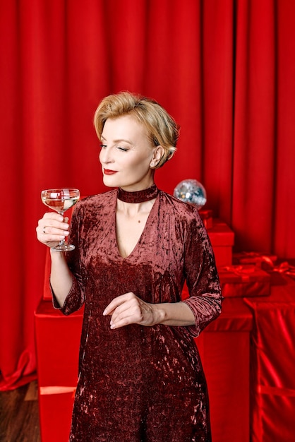 mature stylish elegant woman with glass of sparkling wine with presents on red background.