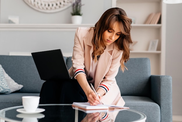 Photo mature successful business woman writing in the notebook using laptop while at home working