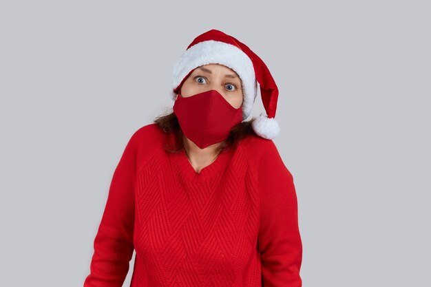 Mature surprised woman in a red protective mask, in a santa hat on a gray background. Christmas, New Year in quarantine.