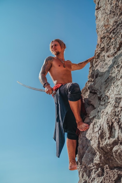 Mature tattooed man holding sword ready to fight and climbing on the rock with background of blue