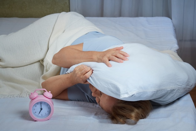 A mature woman covered her head with a pillow, awakened by the ringing alarm clock. The lady is suffering from insomnia.