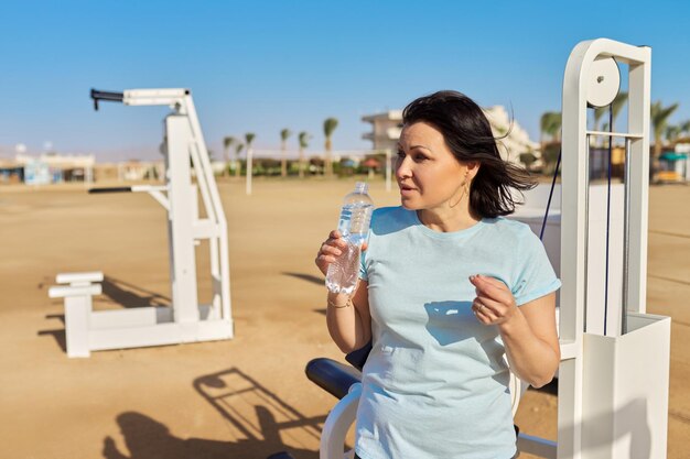 Mature woman doing sports exercise on outdoor simulators with bottle of water